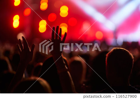 Fans raise hands in excitement at vibrant outdoor music fest, lights illuminate stage, dynamic atmosphere. Energetic crowd enjoys live concert, nightlife entertainment. Excitement fills open event. 116527900