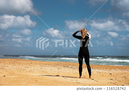 young woman in a wetsuit stands on the beach on the sand against the backdrop of a stormy sea with waves and looks to the side, rear view. Girl in neoprene suit young woman in a wetsuit stands on the beach on the sand against the backdrop of a stormy sea with waves and looks to the side, rear view. Girl in neoprene suit 116528742