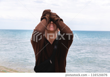 The woman screams with excitement and smiles hysterically, covering her eyes with her hands. A girl in a cardigan at the sea dramatically experiences feelings 116528876