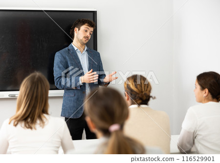 Young male teacher near interactive board, gesturing with hand while consecrating topic of lesson for adults in classroom 116529901