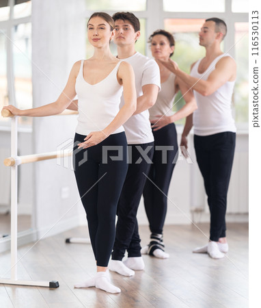 Group of young dancers engaged in ballet in dance studio perfoms exercise near ballet barre, male choreographer helps to coordinate movements correctly Group of young dancers engaged in ballet in dance studio perfoms exercise near ballet barre, male choreographer helps to coordinate movements correctly 116530113
