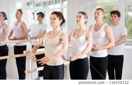 Happy woman in black and white sportswear, participating first position of ballet stance at barre with group in choreography studio 116530132