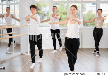 Experienced young female ballet dancer, dressed in white top and black leggings, executing dance moves with focused precision among group of people rehearsing in ballet studio 116530151