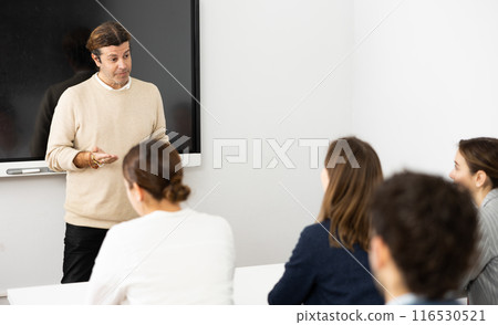 Male trainer near interactive board conducting advanced training courses to office employees sitting at desks in auditory Male trainer near interactive board conducting advanced training courses to office employees sitting at desks in auditory 116530521