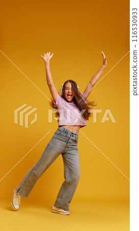 Full-length of pretty young girl in pink t-shirt and grey jeans listening to music in headphones and dancing against yellow studio background 116530933