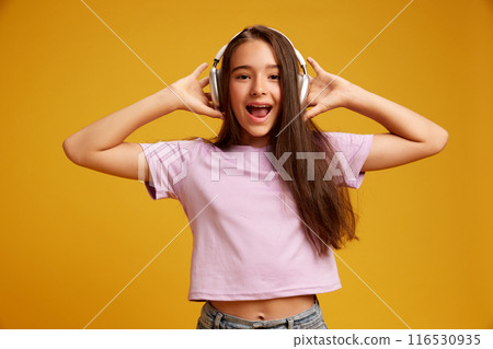 Portrait of beautiful happy teen girl with long brown hair, in pink t-shirt listening to music in headphones and smiling against yellow studio background 116530935