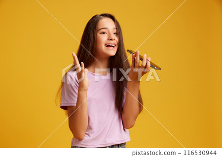Positive and emotional teen girl in pink t-shirt, emotionally recording voice message, talking on mobile phone against yellow studio background 116530944