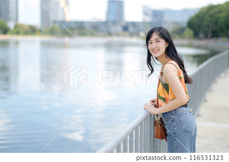 Urban charm meets natural bliss as a stylish woman in orange shirt enjoys a sunny day by the lake. Urban charm meets natural bliss as a stylish woman in orange shirt enjoys a sunny day by the lake. 116531323