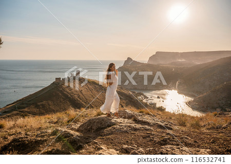 woman stands on a rocky hill overlooking a body of water. She is wearing a white dress and she is in a state of joy or celebration. Concept of freedom and happiness. woman stands on a rocky hill overlooking a body of water. She is wearing a white dress and she is in a state of joy or celebration. Concept of freedom and happiness. 116532741