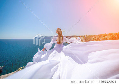 woman sea white dress. Blonde with long hair on a sunny seashore in a white flowing dress, rear view, silk fabric waving in the wind. Against the backdrop of the blue sky and mountains on the seashore 116532804