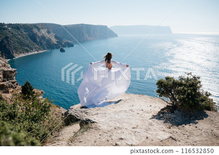 woman white dress stands on a rocky cliff overlooking the ocean. The scene is serene and peaceful, with the woman's dress billowing in the wind. The ocean in the background is calm and blue. woman white dress stands on a rocky cliff overlooking the ocean. The scene is serene and peaceful, with the woman's dress billowing in the wind. The ocean in the background is calm and blue. 116532850