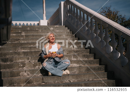 A woman sits on a set of stairs, holding a cup of coffee. She is enjoying her coffee and taking in the view from the top of the stairs. 116532935