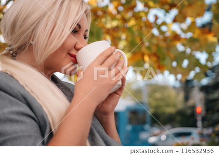 A blonde woman is drinking coffee while sitting under a tree. The scene is peaceful and relaxing, with the woman enjoying her coffee and the natural surroundings. 116532936