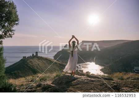woman stands on a rocky hill overlooking a body of water. She is wearing a white dress and she is in a state of joy or celebration. Concept of freedom and happiness. 116532953