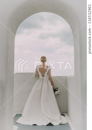 A bride stands in front of an archway, holding a bouquet of flowers. The archway is white and the sky is cloudy. The bride is wearing a white dress and her hair is styled in a bun. A bride stands in front of an archway, holding a bouquet of flowers. The archway is white and the sky is cloudy. The bride is wearing a white dress and her hair is styled in a bun. 116532961