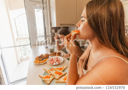 mom and daughter sitting at a table with a variety of food, including sandwiches, fruit, and pastries. They are smiling and enjoying their meal together. 116532970