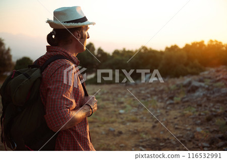 A man wearing a hat and backpack hikes through a scenic landscape during sunset 116532991