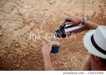 Person pouring hot beverage from thermos into cup while sitting on dry ground outdoors Person pouring hot beverage from thermos into cup while sitting on dry ground outdoors 116532998