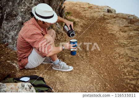 Man sitting on a rocky path and pouring coffee from a thermos into a cup during an outdoor adventure Man sitting on a rocky path and pouring coffee from a thermos into a cup during an outdoor adventure 116533011