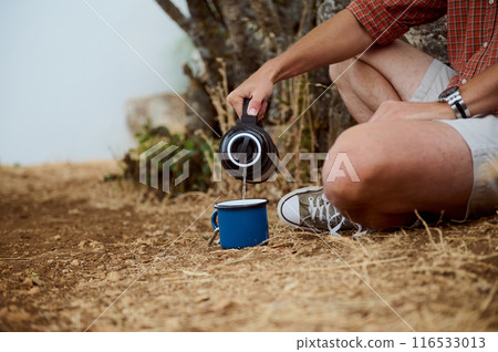 Man pouring hot drink from thermos into a blue mug while sitting outdoors in nature 116533013