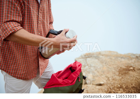 Man preparing hot drink during hike on mountain trail, holding thermos near backpack 116533035