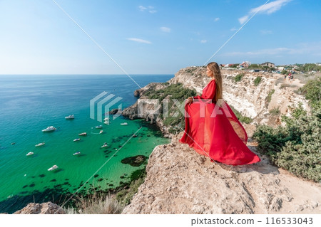 Red Dress Woman sea Cliff. A beautiful woman in a red dress and white swimsuit poses on a cliff overlooking the sea on a sunny day. Boats and yachts dot the background. Red Dress Woman sea Cliff. A beautiful woman in a red dress and white swimsuit poses on a cliff overlooking the sea on a sunny day. Boats and yachts dot the background. 116533043