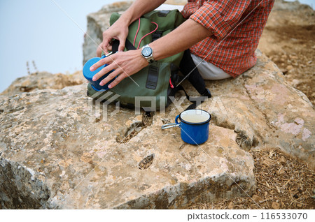 Hiker preparing a drink while sitting on rocky terrain with backpack and blue enamel mug Hiker preparing a drink while sitting on rocky terrain with backpack and blue enamel mug 116533070