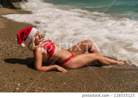 Female beach Santa hat wave coast. beach relaxation seaside. A woman in a red swimsuit enjoying her time on the beach, lying on the sand and being covered by a wave. 116533125