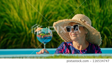 Elderly woman with a delighted expression, wearing a sun hat and sipping a cold drink while lounging in a pool on a bright day Elderly woman with a delighted expression, wearing a sun hat and sipping a cold drink while lounging in a pool on a bright day 116534747