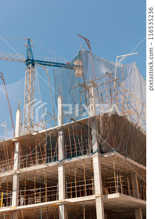 Bustling construction site with a towering crane and protective netting under a clear blue sky, perfect for use in articles about urban development, construction safety, and real estate projects. 116535236