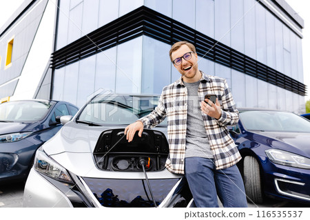 A young man uses a mobile phone while leaning on his electric car while the car is charging A young man uses a mobile phone while leaning on his electric car while the car is charging 116535537
