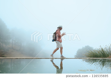 Traveler walking along a misty path by water, using smartphone, wearing hat and backpack Traveler walking along a misty path by water, using smartphone, wearing hat and backpack 116535695