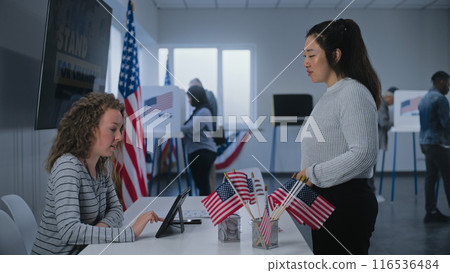 Female polling officer consults Asian woman and gives ballot paper for voting Female polling officer consults Asian woman and gives ballot paper for voting 116536484