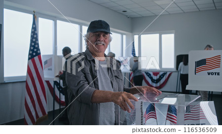 Elderly Caucasian man puts ballot paper in box, then smiles and looks at camera Elderly Caucasian man puts ballot paper in box, then smiles and looks at camera 116536509