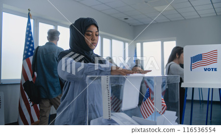 Muslim woman in hijab puts ballot paper in box at polling station 116536554