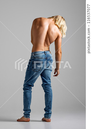 Back view of young man with long blonde hair posing shirtless against grey studio background. Model in denim jeans. Back view of young man with long blonde hair posing shirtless against grey studio background. Model in denim jeans. 116537077