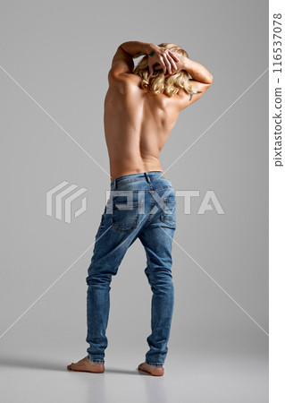Rear view photo of young man with long blonde hair, posing dressed denim jeans and holding hands behind head against grey studio background. Rear view photo of young man with long blonde hair, posing dressed denim jeans and holding hands behind head against grey studio background. 116537078