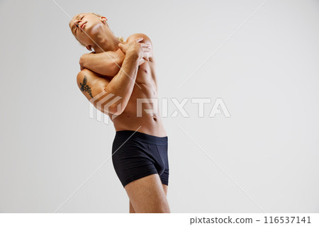 Athletic man wearing black boxer briefs poses with his arms wrapped around his upper body and head leaned back against white studio background. 116537141