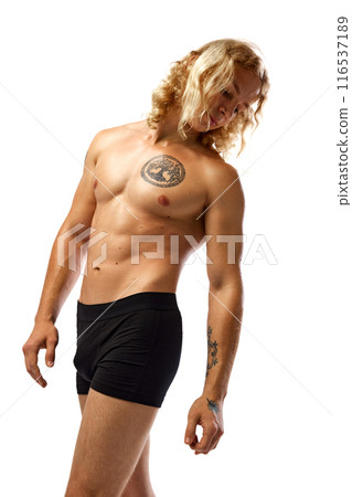 Muscular man with long, curly blonde hair stands in black boxer briefs, showing off his chest tattoo and arm tattoos. against white studio background. Muscular man with long, curly blonde hair stands in black boxer briefs, showing off his chest tattoo and arm tattoos. against white studio background. 116537189