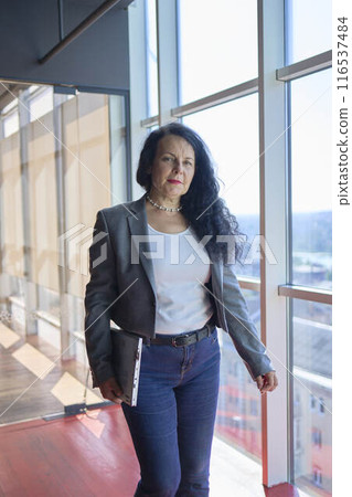 A 57-year-old woman walks with a laptop to her glass office in a modern workspace 116537484