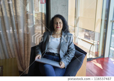 A 57-year-old woman sits with a laptop in a chair in a modern workspace 116537487