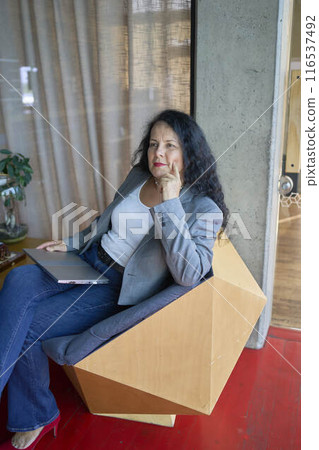 A 57-year-old woman sits with a laptop in a chair in a modern workspace 116537492