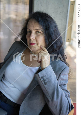 A 57-year-old woman sits with a laptop in a chair in a modern workspace 116537494