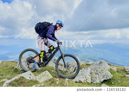 Cyclist man riding electric mountain bike outdoors. Male tourist biking along grassy trail in the mountains, wearing helmet and backpack. Concept of sport, active leisure and nature. 116538182