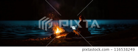 晚上，一名婦女在湖邊篝火前使用筆記型電腦工作的後視圖 116538249