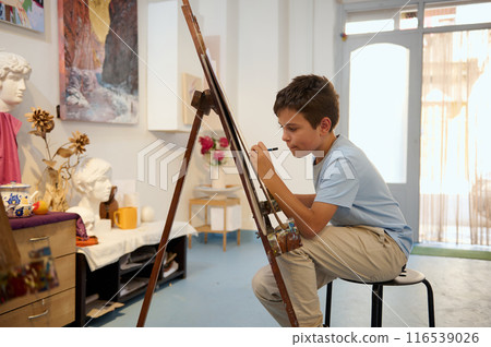 Young boy having an art class, drawing and painting on an easel with concentration in a creative workshop setting 116539026