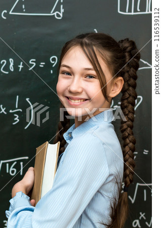A high school schoolgirl stands near the blackboard with a textbook in her hands, portrait of a teenage girl at school A high school schoolgirl stands near the blackboard with a textbook in her hands, portrait of a teenage girl at school 116539112