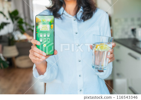 Healthy habit to drink water. Blurred no face woman with glass of pure water with lemon holding phone with active mobile app to control body hydration, Tracking water balance. Selective focus. 116543346