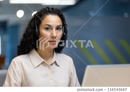 Focused professional woman with curly hair and headset working at desk in office environment. Modern workplace with blue and green decor. Ideal image depicting customer service, support, and Focused professional woman with curly hair and headset working at desk in office environment. Modern workplace with blue and green decor. Ideal image depicting customer service, support, and 116543687