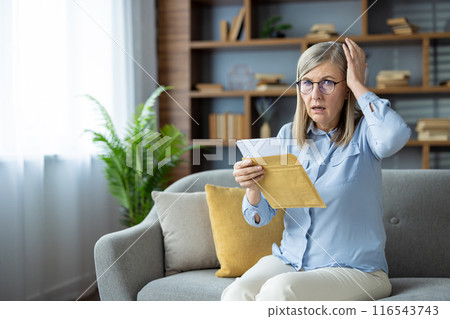 Shocked elderly woman receiving unexpected news while reading letter on sofa at home. Senior lady with glasses and hand on head expresses surprise or worry. 116543743